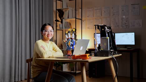 teen asian girl with a laptop smiling and crossing her arms to the camera while working about a cyborg hand at home