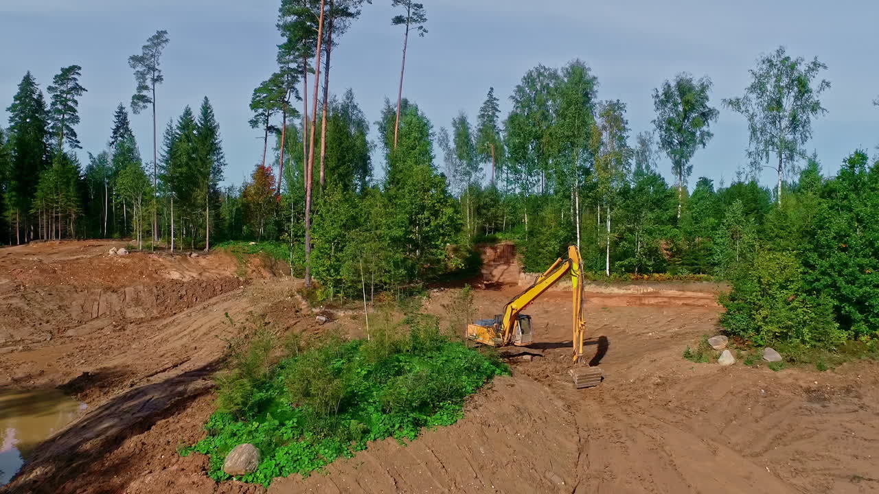 Tung gravemaskine, der arbejder ved en dam i et skovområde, set fra luften.