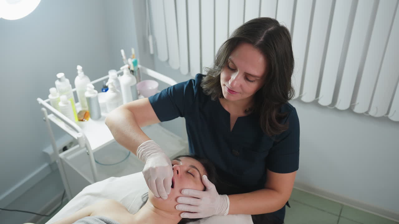 High angle of spa guest lying on massage chair opens mouth and closes eyes gently as professional wearing safety gloves dips fingernail inside mouth and uses other hand to massage cheeks