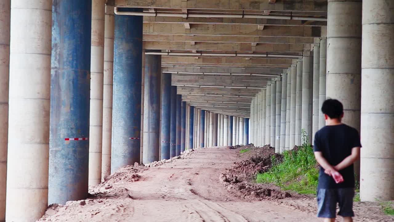 un joven asiático soltero caminando bajo un puente con tierra y plantas en el fondo