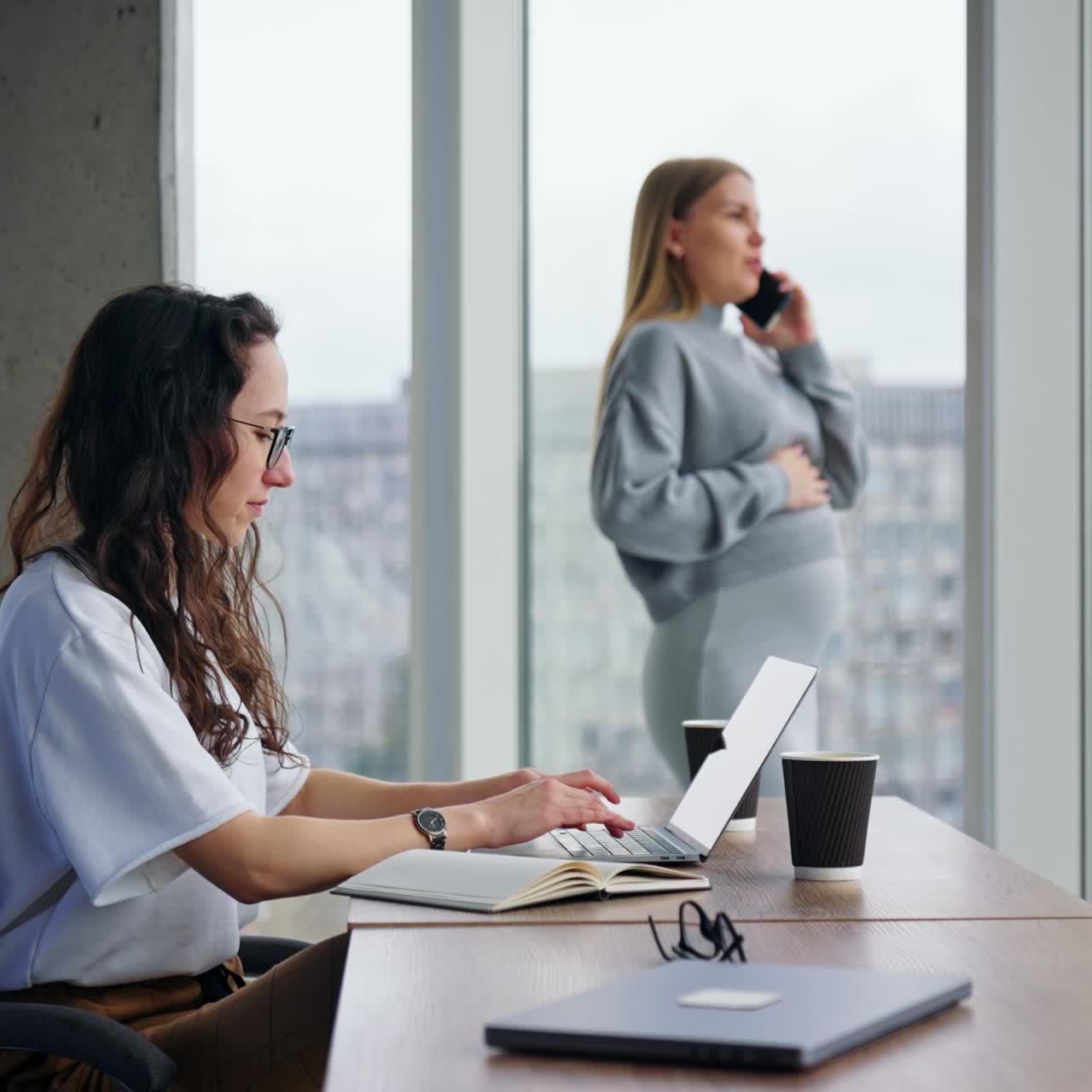 Signora bruna, seria e concentrata sul lavoro, lavora al laptop. Signora incinta parla al telefono in piedi e guarda la finestra sullo sfondo