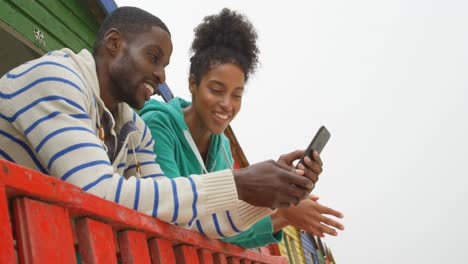 Vista-Lateral-De-Una-Joven-Pareja-Negra-Usando-Un-Teléfono-Móvil-Mientras-Se-Apoya-En-Una-Barandilla-En-Una-Cabaña-En-La-Playa-4k