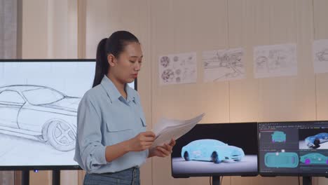 mujer asiática diseñadora de automóviles mirando papeles de boceto de diseño de automóviles en sus manos y caminando por el estudio con la escultura de arcilla de automóviles