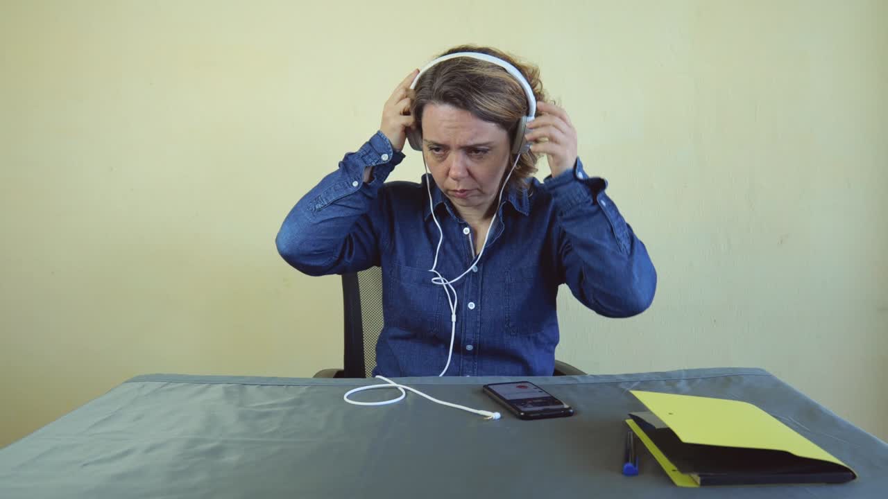 An adult woman in a denim shirt takes off white large headphones. A woman is talking on a mobile phone while sitting at a table
