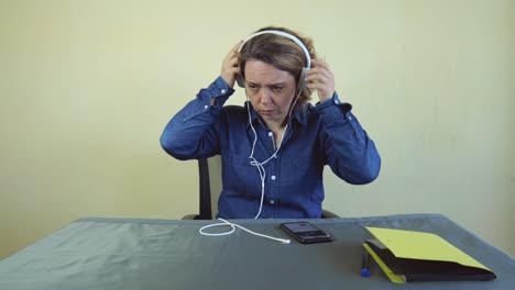 an adult woman in a denim shirt takes off white large headphones. a woman is talking on a mobile phone while sitting at a table