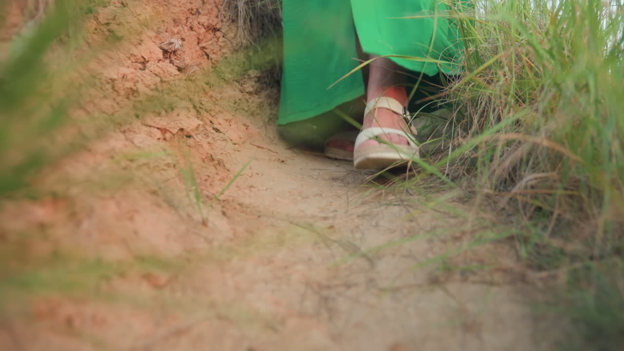 Lower angle closeup showing feet of woman in green gown cautiously stepping down sandy narrow hill path bordered by dry roots and fresh grass