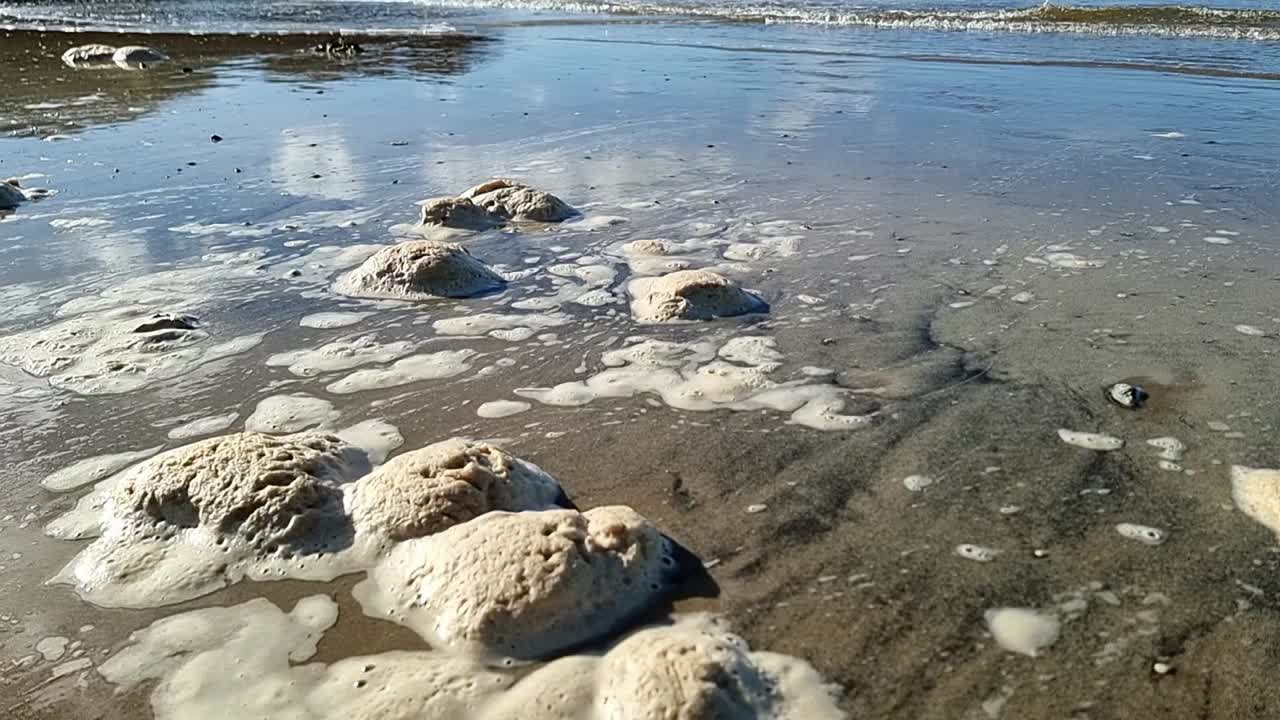 Schiuma di mare sporca che si muove lentamente attraverso la spiaggia sabbiosa di Anglesey, in Galles, con le onde della marea