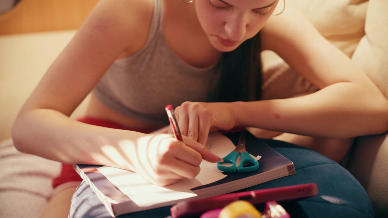 Studentessa delle scuole superiori profondamente concentrata e preoccupata che prende appunti segreti e piccoli bigliettini per copiare dal suo telefono cellulare, preparandosi per un esame mentre è seduta su un divano sotto una calda illuminazione interna.