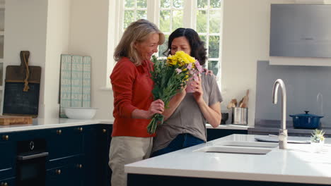 woman surprising same sex mature female partner with flowers  in kitchen at home