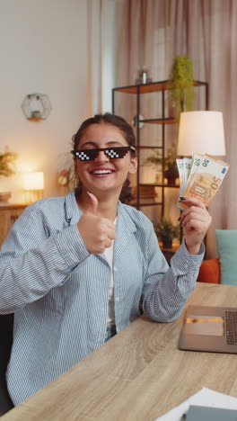 young woman freelancer putting on party glasses holding fan of cash sitting at home office table