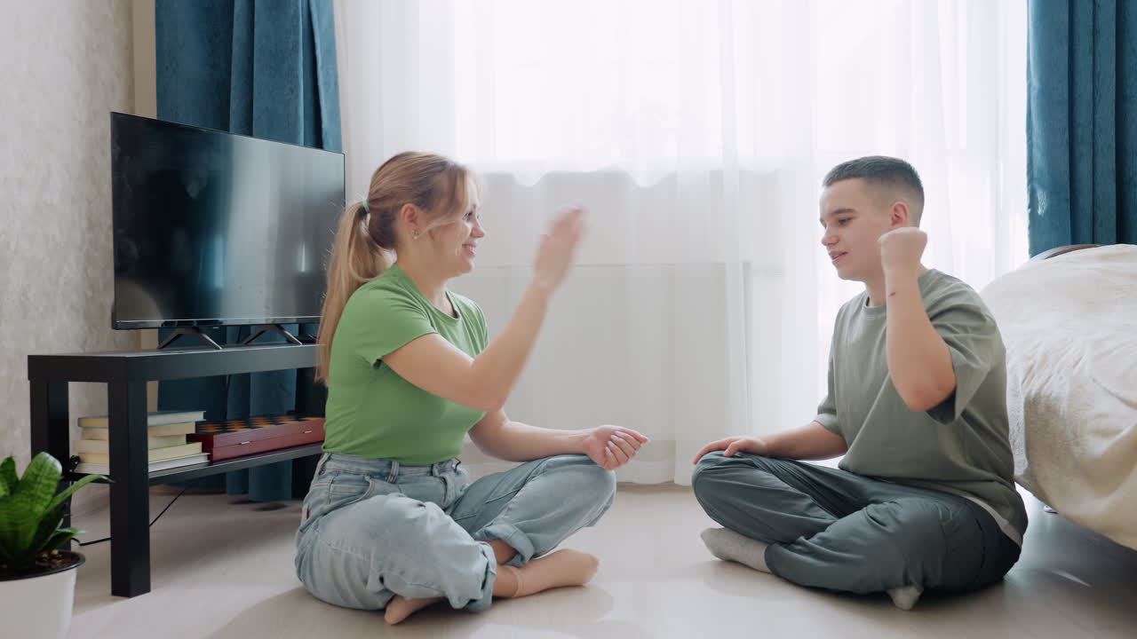 Mother and son enjoy playful hand game while sitting barefoot on floor near bed and television in cozy bright room, surrounded by books, indoor plant, and sunlight through sheer curtains