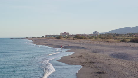 Beautiful-mediterranean-coast-with-people-walking-along-the-beach-with-their-dogs,-with-buildings-and-mountains-on-the-horizon,-wide-shot