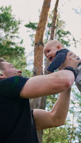 handsome muscular caucasian man tossing his little baby boy. spending time with child in nature. low angle view. pine trees at backdrop. vertical video