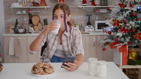 chica feliz disfrutando de las vacaciones de invierno sentada en la mesa en la cocina decorada de navidad navegando en su teléfono inteligente