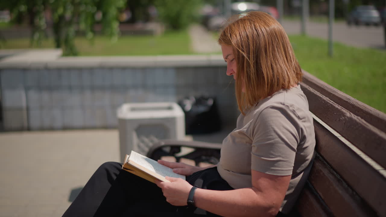 Woman sitting on wooden bench reading book outdoors on sunny day, relaxed expression and natural posture, blurred car movement in background