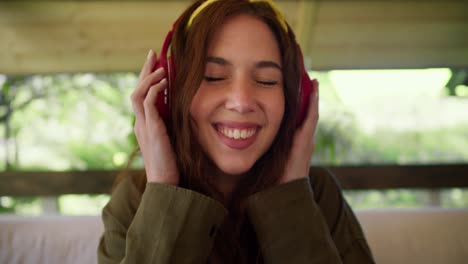 a brunette girl in a green sweater with a yellow bandage on her head puts on red wireless headphones listen to music and shakes her head while sitting on a sofa in a gazebo in nature