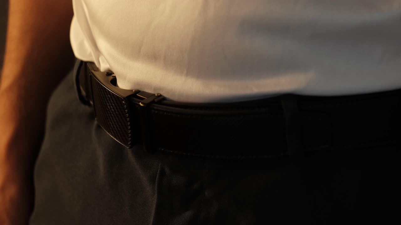 Close-up static shot of man wearing and adjusting ratchet belt over dress pants and white shirt