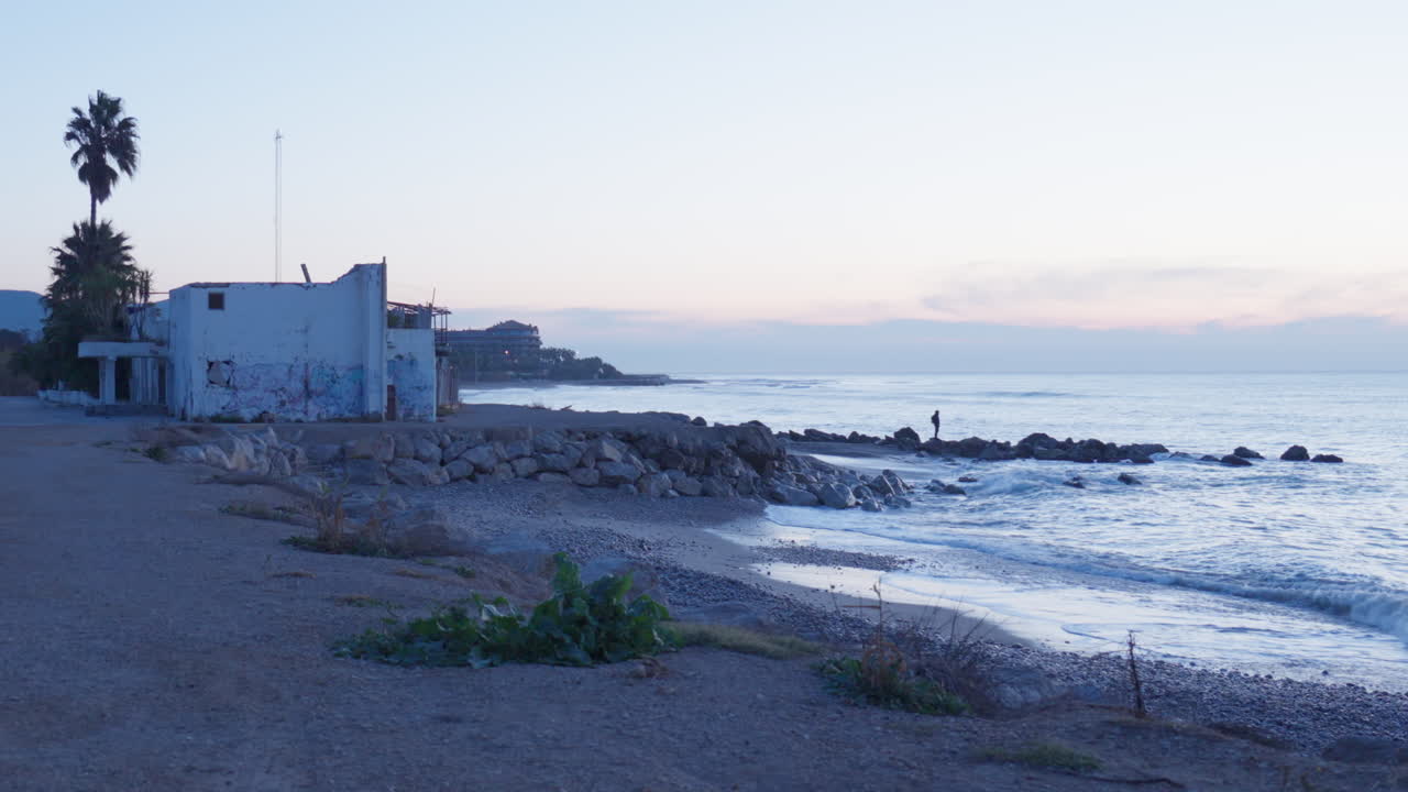 한 남자가 비치 하우스의 바위가 많은 부두에 서서 이른 아침 햇살 아래 밀려오는 파도를 바라보고 있습니다.