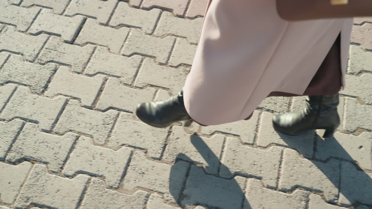 Woman walking on patterned pavement pink coat and black heeled boots, closeup of confident stride and swinging handbag, executive commuter hurrying between meetings, shopper pausing to adjust