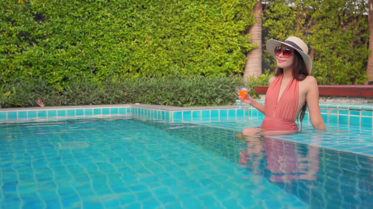 Young woman holding a tropical drink in one hand as she lounges on the step to the shallow end of a swimming pool. Copy space
