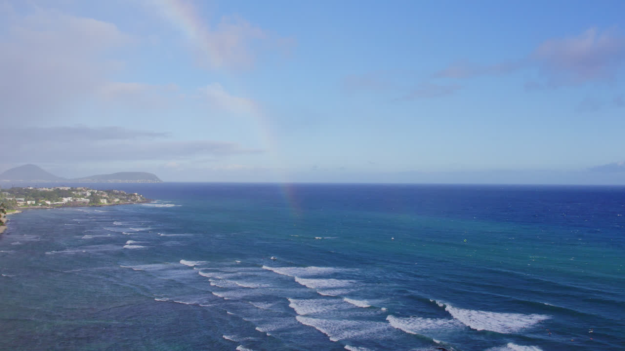las imágenes de drones capturan un arco iris en la niebla del océano a lo largo de la costa de oahu en las islas hawaianas con montañas volcánicas en el horizonte y olas blancas que rodan sobre la costa.