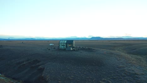 Orbital-shot-of-a-crystal-glass-cabin-on-a-hill-in-a-black-and-dark-landscape,-Iceland-destination