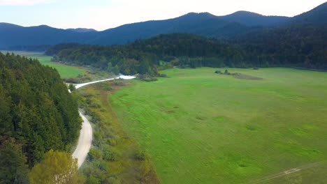 Aerial-Over-Road-Next-To-Open-Green-Field-With-Forested-Hills-In-The-Distance