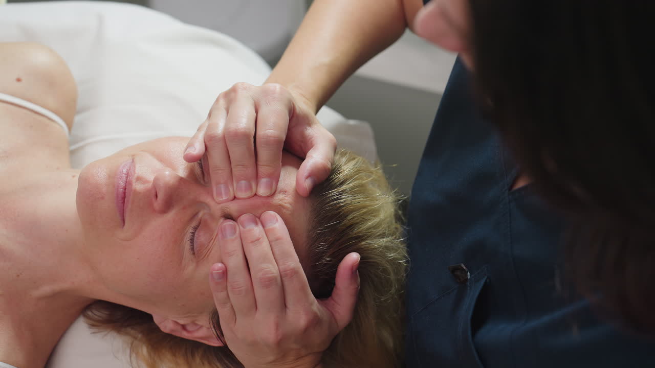 Cosmetologist hands lift forehead skin of woman lying relaxed on massage table, therapist glides fingers to smooth tension as client closes eyes, releases shoulders in serene spa environment