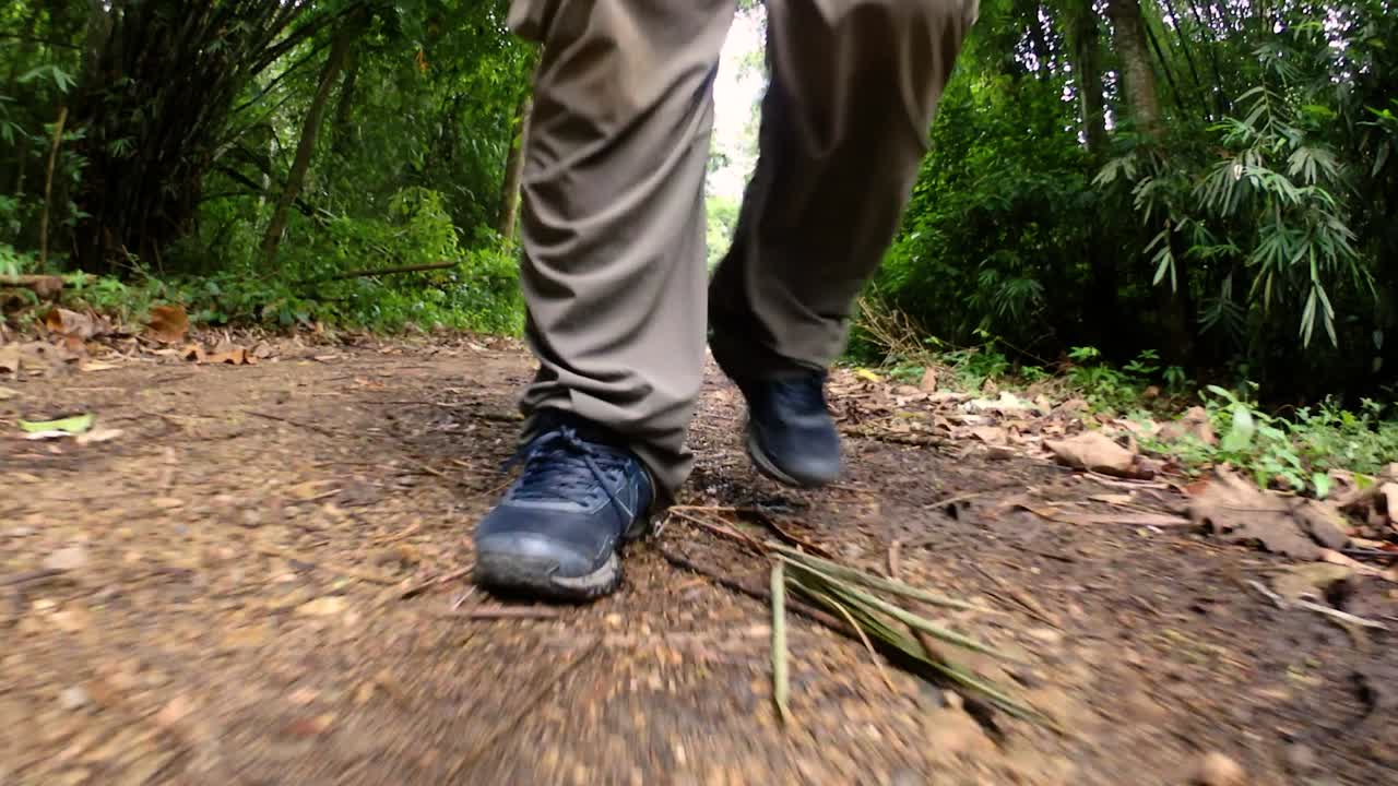 un hombre caminando en la jungla, la cámara enfocada en sus piernas, mostrándolo dando un paso adelante en el camino de tierra con sus zapatos enmarcados