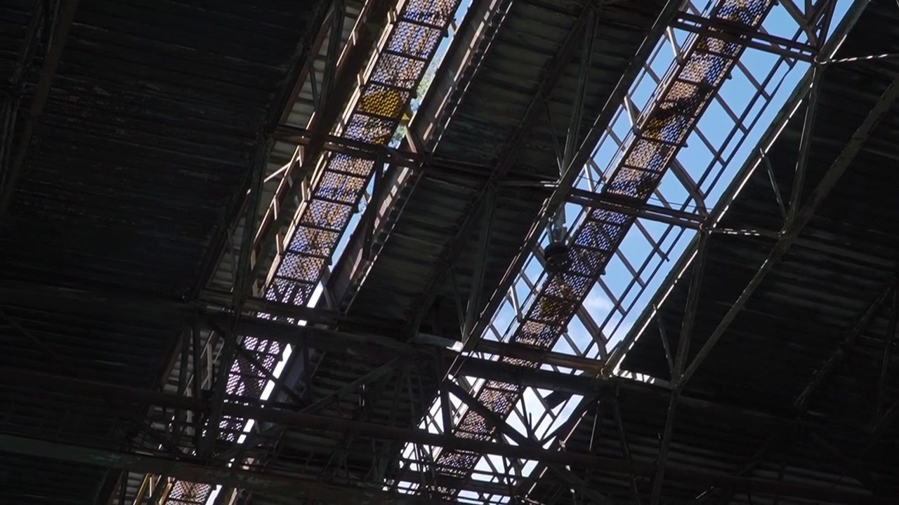 La telecamera cattura intricati giochi di luce solare che filtrano attraverso un tetto industriale, rivelando il cielo azzurro oltre. Ideale per studi architettonici o progetti a tema industriale