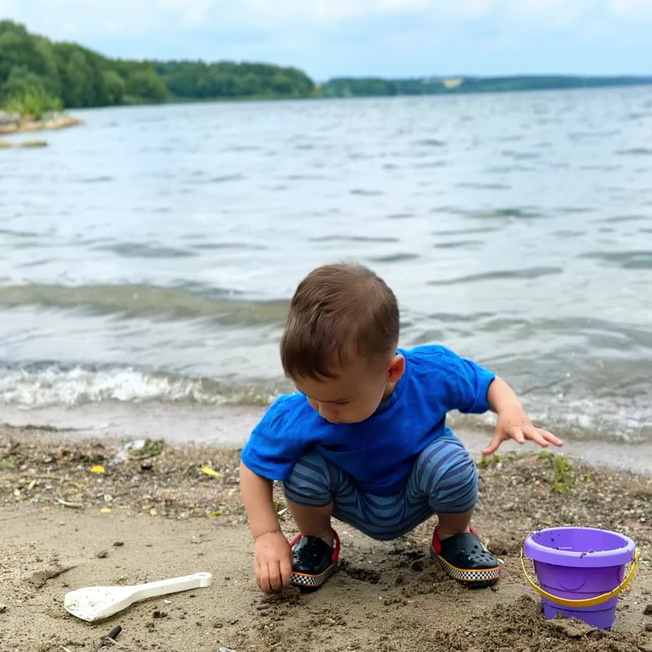 Little toddler in blue t-shirt plays at the river bank. Child takes his toy shovel and walks to the water