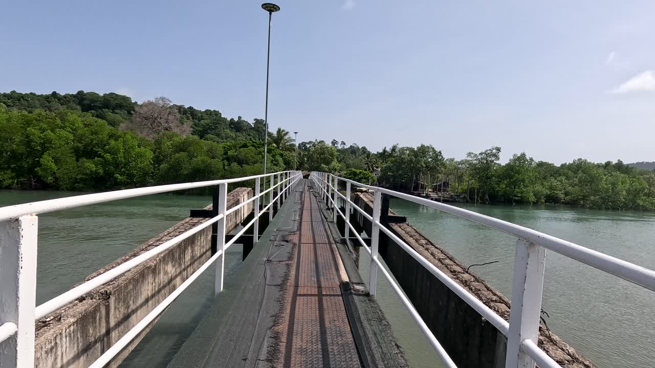 Point-of-view walk along a narrow bridge above a mangrove-lined river, under bright daylight with steady camera movement and lush green surroundings