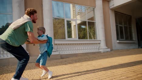 Happy-little-blond-boy-in-blue-clothes-and-with-a-blue-backpack-runs-to-his-dad-in-a-green-T-shirt-who-takes-him-in-his-arms-and-throws-him-up.-Happy-meeting-of-father-and-son-after-boy's-school-activities