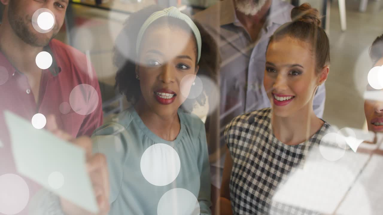 Lead woman pointing and explaining business plan on glass board, tablet entering, team leaning in