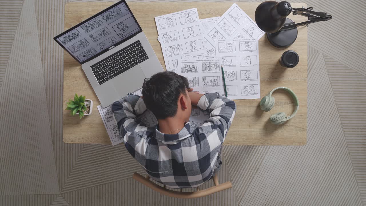 vista superior de un artista masculino asiático cansado durmiendo mientras dibuja un guión gráfico en la mesa con una computadora portátil y auriculares en el estudio