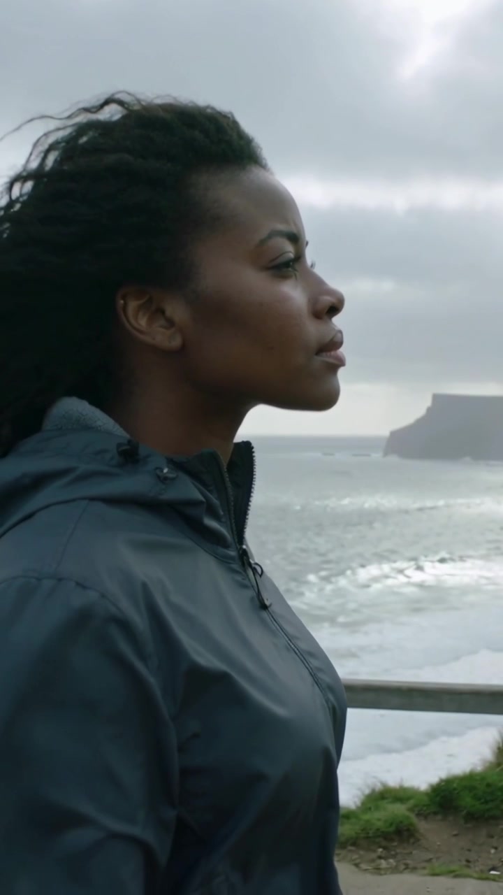 Vertical video: Standing woman wind blowing hair by cliff railing in waterproof coat, reflecting