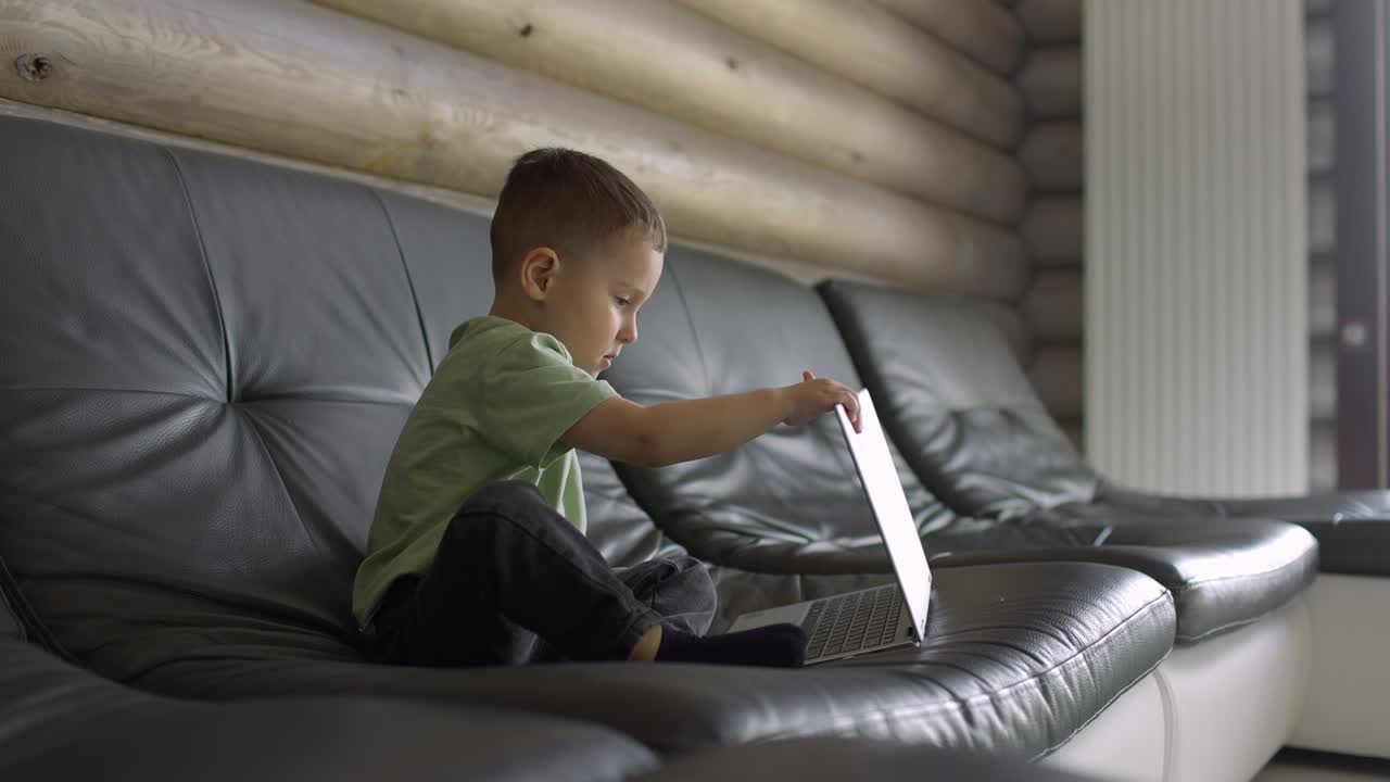 Serious and focused boy covers his laptop lid while sitting on the sofa in daylight