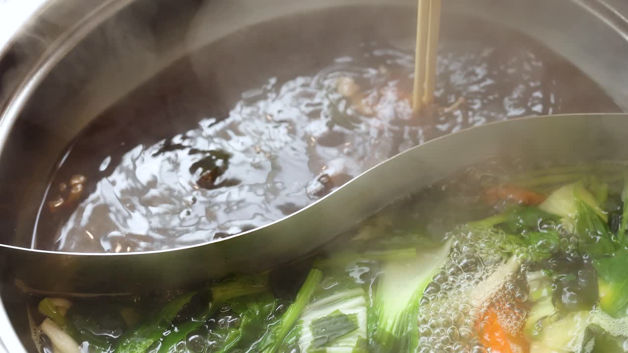 Chopsticks lift cooked beef from steaming dual broth hotpot with vegetables, natural lighting, close-up
