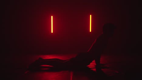 man sitting in kneeled pose does yoga spine stretch in dark room, red lights casting shadow silhouette