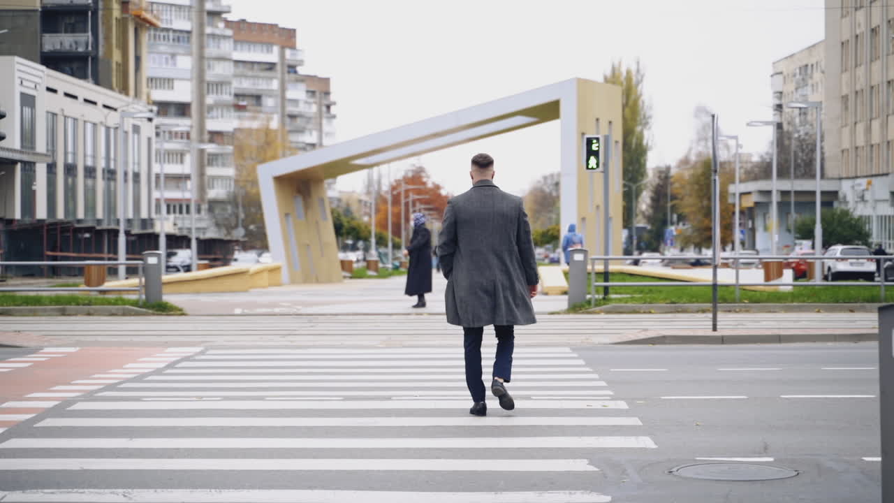 Backside view of a young man in the city. Back of a man in warm coat crossing the road on the green light of the traffic lights. Slow motion.