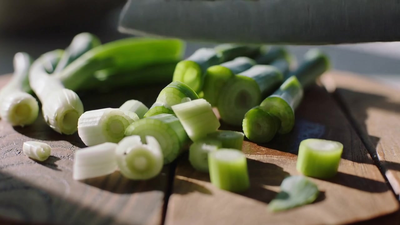 Coltello che affetta cipollotti su tagliere di legno rotondo per cucinare, con ombre.