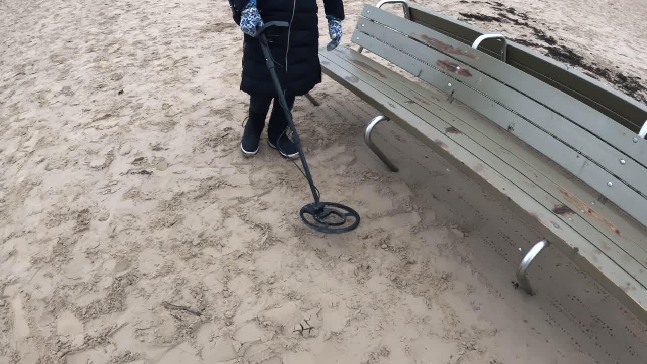Person som skannar sanden runt en sliten strandbänk med en metalldetektor och söker efter tappade föremål nära en sittplats vid kusten