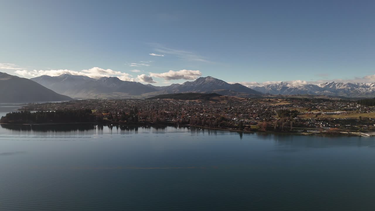 Wanaka town and lake during golden sunrise in New Zealand. Aerial wide shot. Mountain range in blue sky with clouds. Panorama view. Peaceful city on island
