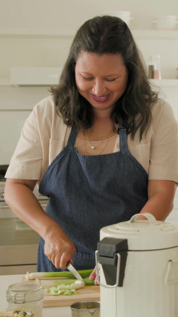 Vertical video: Lifting white cooker lid, woman in blue apron leading at island, chopping scallions