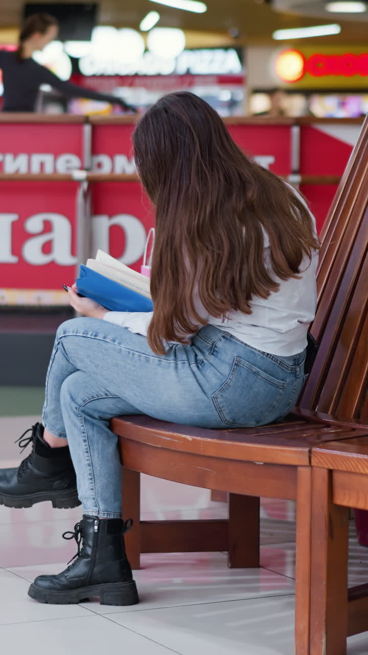 vista trasera de una mujer sentada en un banco de madera, con las piernas cruzadas, leyendo un libro, el cabello largo fluyendo, compradores visibles en el fondo, entorno vibrante del centro comercial