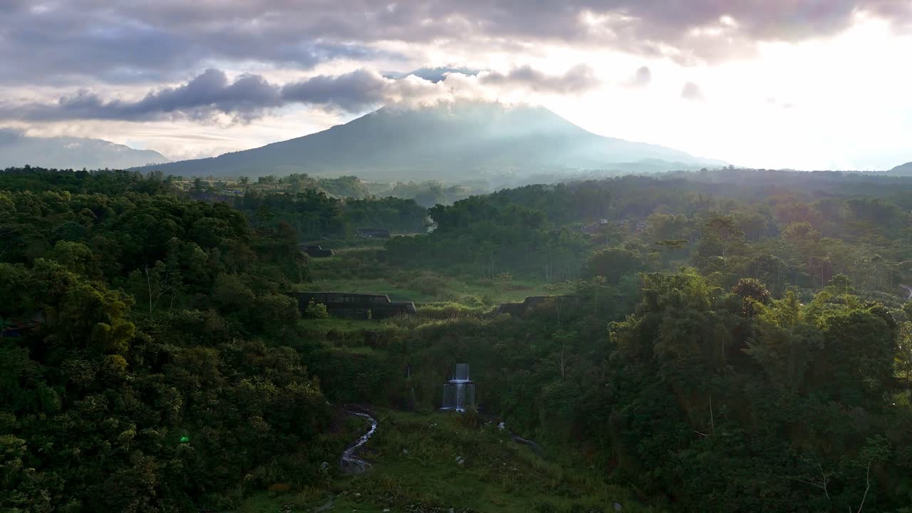 Aerial view of a waterfall in the jungle with Indonesia's Merapi volcano in the background.