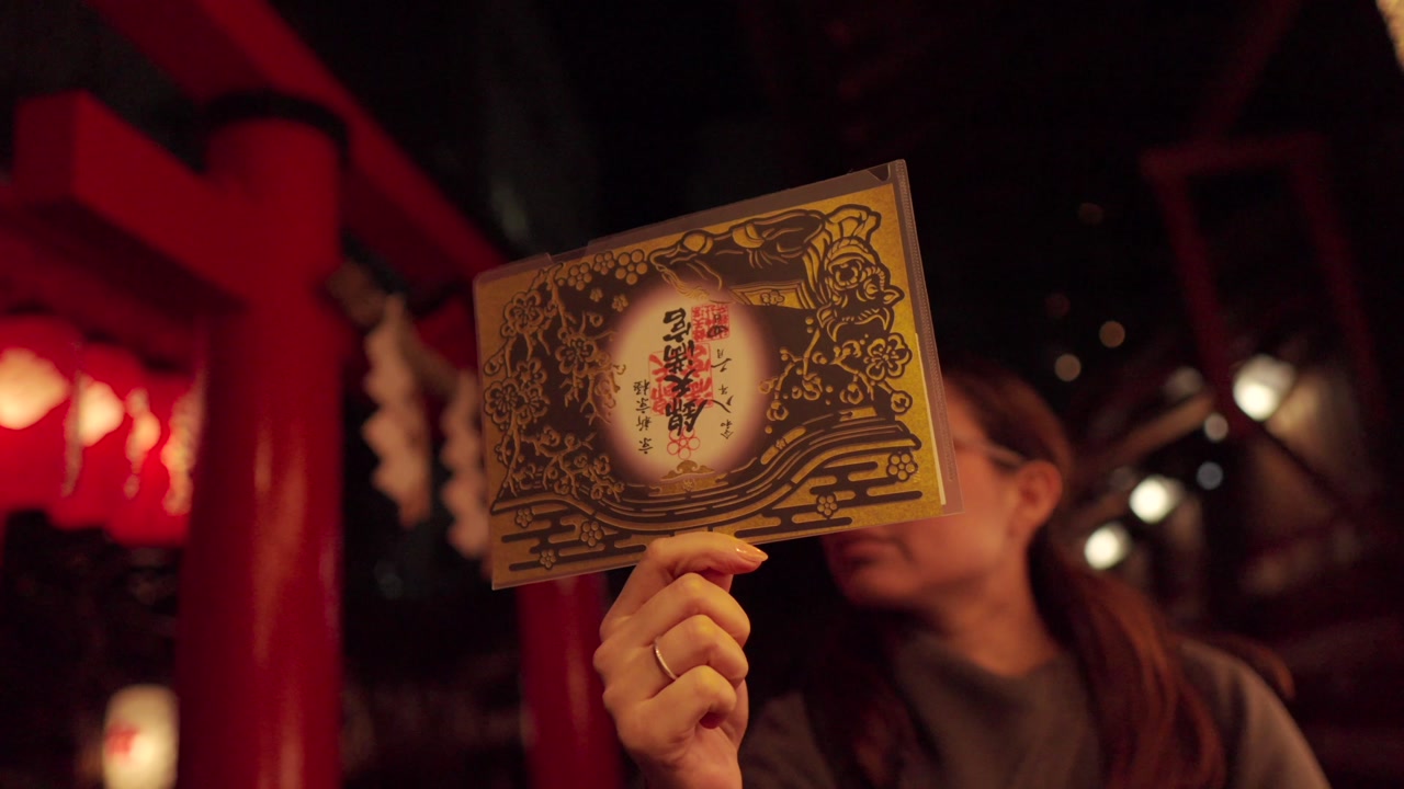 A young Latina tourist holds up a Goshuin stamp in front of traditional lanterns at Nishiki Tenmangu Shrine in Kyoto, Japan, at night. The camera orbits around her, capturing the sacred atmosphere.