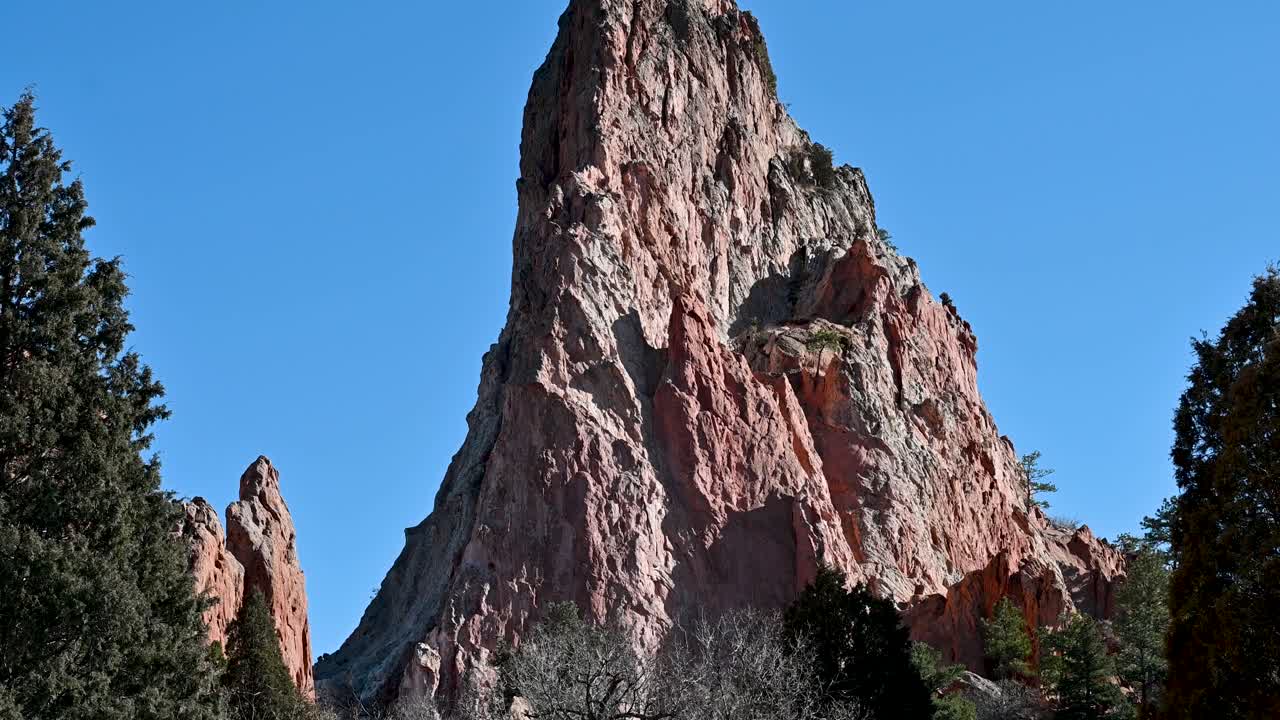 Drone shot of towering red sandstone spire at Garden of the Gods, framed by trees and vibrant blue sky in Colorado Springs