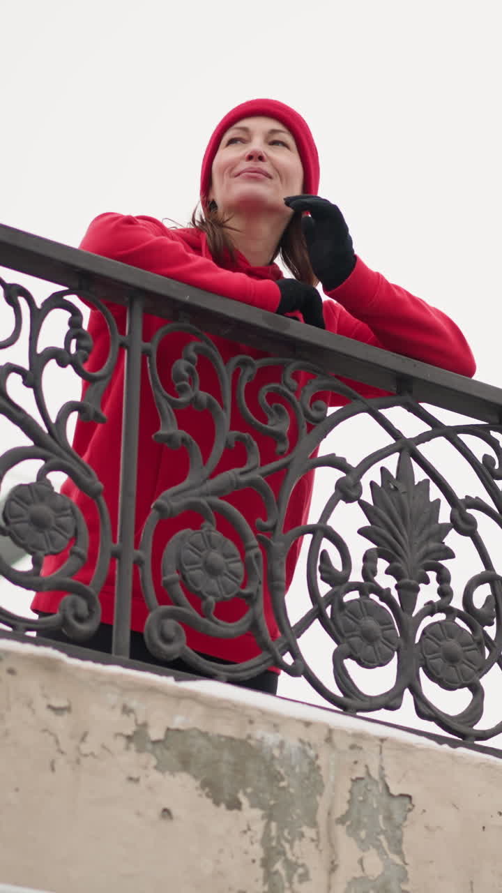 Woman in red beanie and hoodie rests hands on intricate iron railing under bright winter sky, smiling with serene expression and placing right hand on head while gazing into distance