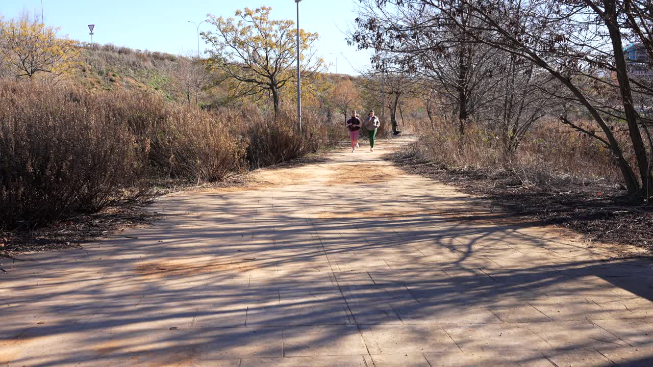 Two women running outdoors on a sunny path; one tired, the other encouraging her
